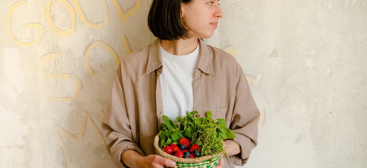 Woman standing with a basket of fresh greens and berries against a rustic wall background.