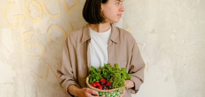 Woman standing with a basket of fresh greens and berries against a rustic wall background.